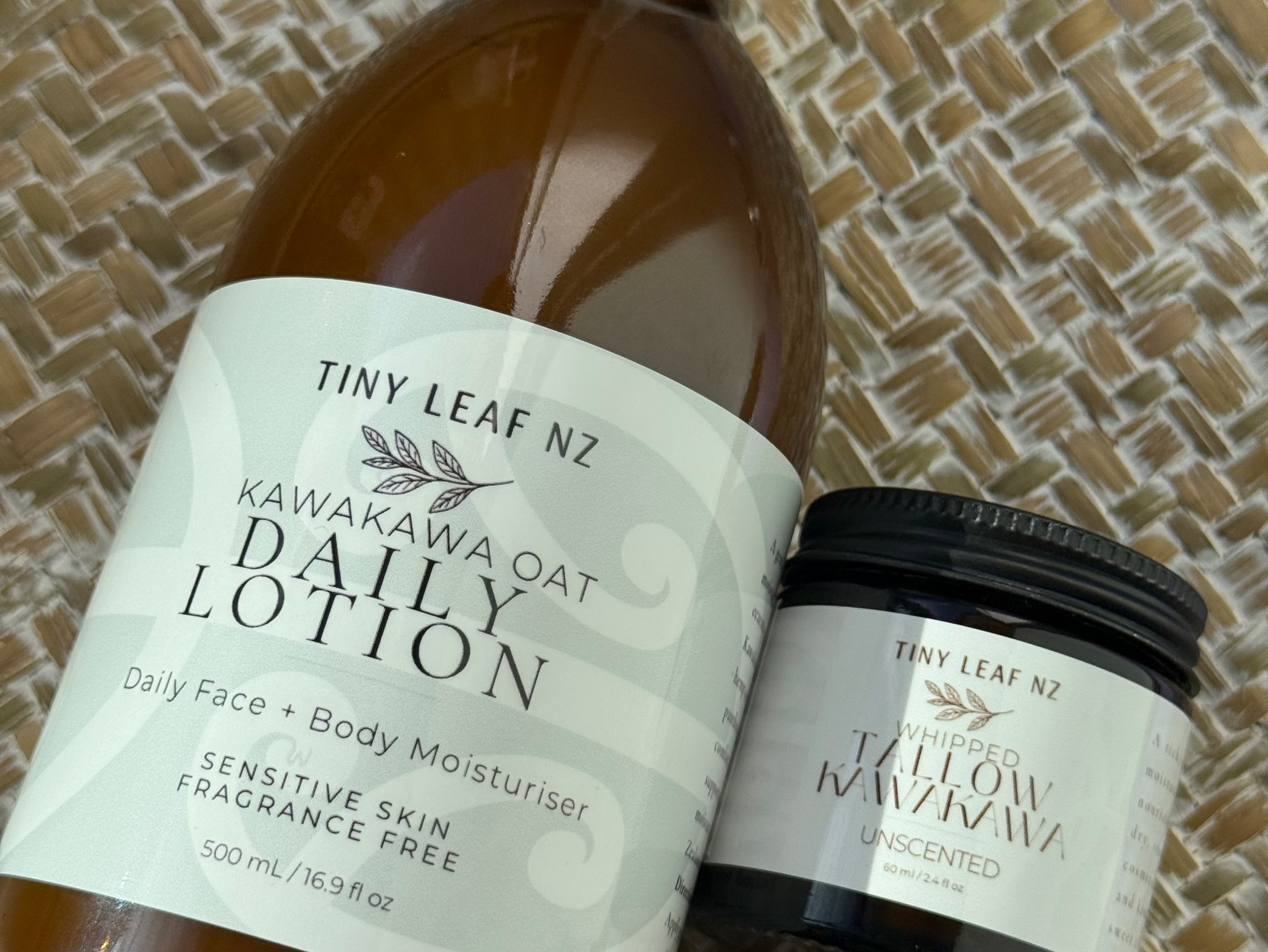 A bottle of Tiny Leaf NZ Kamakawa Daily Lotion with a white dropper cap and a small jar of the same lotion with a black lid, both with white labels, displayed on a woven straw mat.
