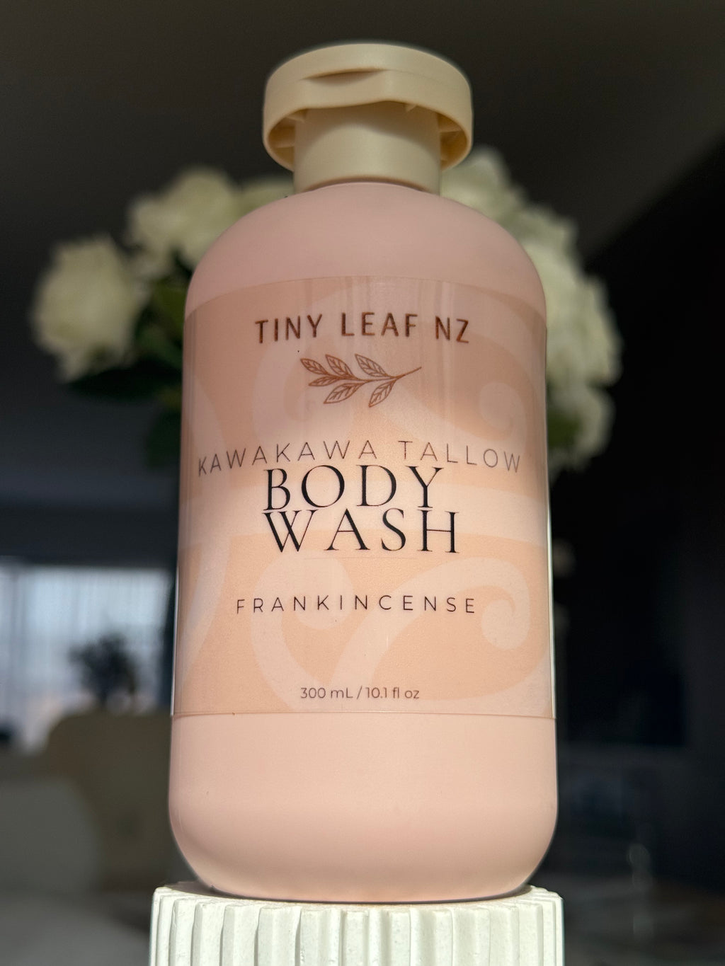 A bottle of Tiny Leaf NZ Kawakawa Tallow Body Wash in Frankincense scent, featuring a pale pink bottle and cap, displayed on a white pedestal with a blurred background of white flowers.