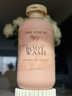 A bottle of Tiny Leaf NZ Kawakawa Tallow Body Wash in Frankincense scent, featuring a pale pink bottle and cap, displayed on a white pedestal with a blurred background of white flowers.