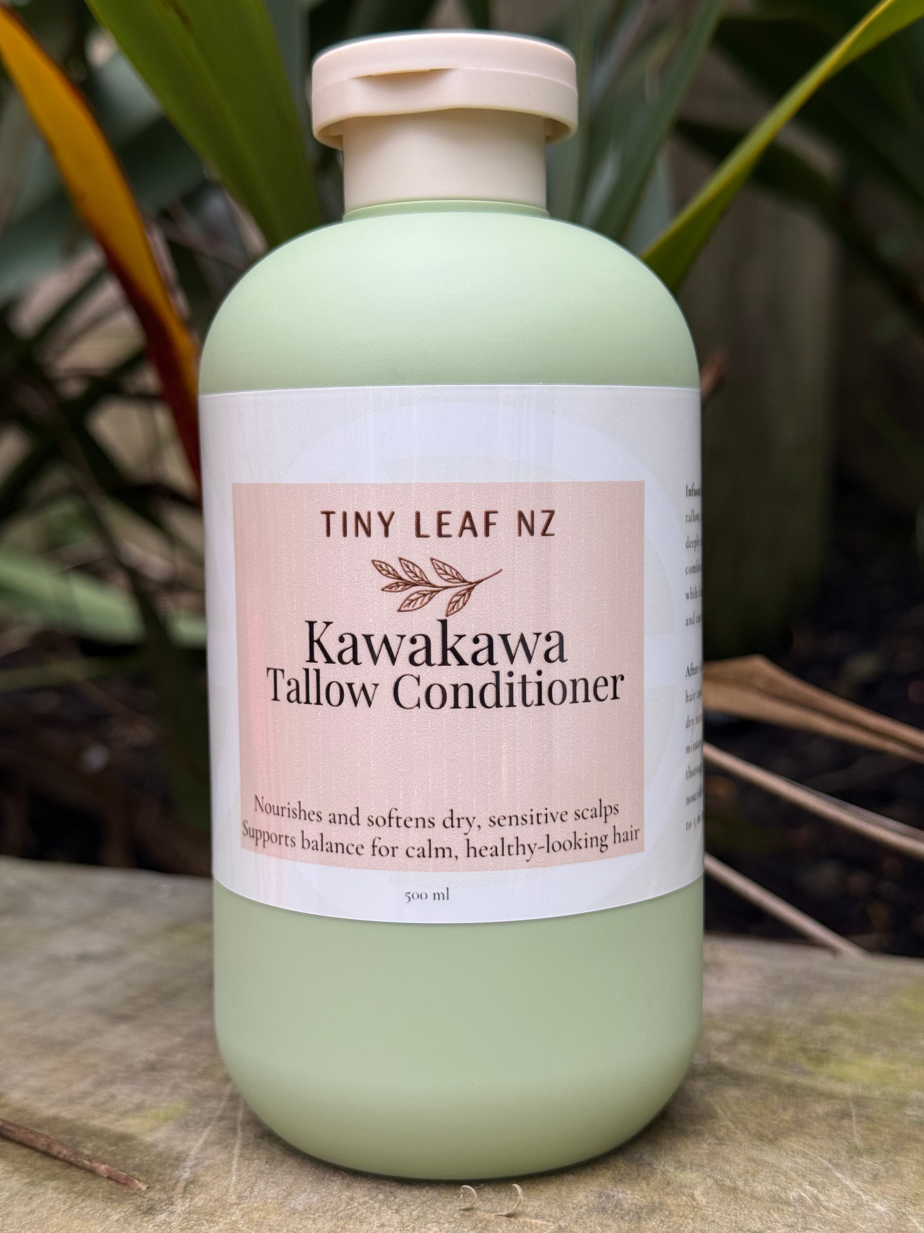 A 500 ml bottle of Tiny Leaf NZ Kawakawa Tallow Conditioner, featuring a pale sage green body and a white label with a pinkish-beige accent, set against a natural background of green leaves.