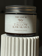 A small amber glass jar candle with a silver metal lid and a cream label reading 'TINY LEAF NZ WHIPPED TALLOW KAWAKAVA FRANKINSCENCE', displayed on a white ribbed pedestal against a dark background.