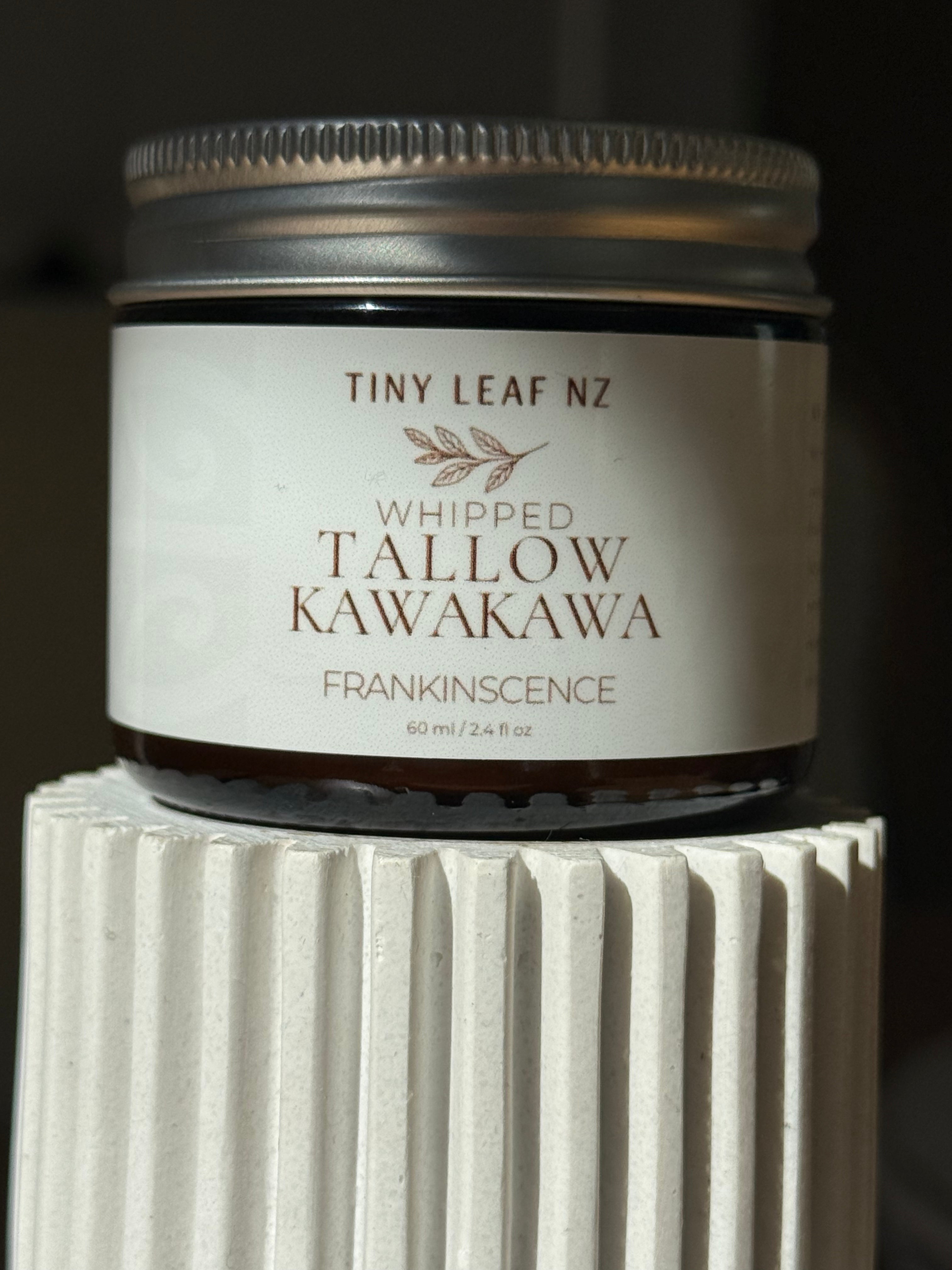 A small amber glass jar candle with a silver metal lid and a cream label reading 'TINY LEAF NZ WHIPPED TALLOW KAWAKAVA FRANKINSCENCE', displayed on a white ribbed pedestal against a dark background.