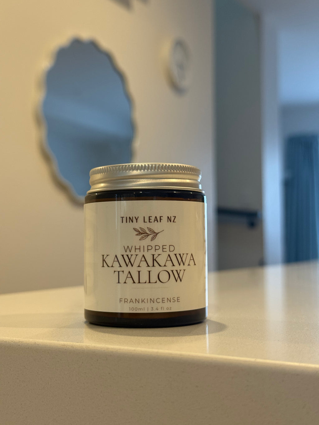 A jar of Tiny Leaf Nz Whipped Kawakawa Tallow Frankincense wax melt sits on a light countertop in a home setting with a blurred background.