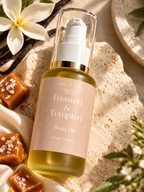 A bottle of Tiny Leaf's 'Toasted & Tempted' body oil, filled with golden-yellow oil and featuring a pale pink label with gold lettering, styled on a textured beige stone surface with caramel squares, a white flower, and vanilla beans.