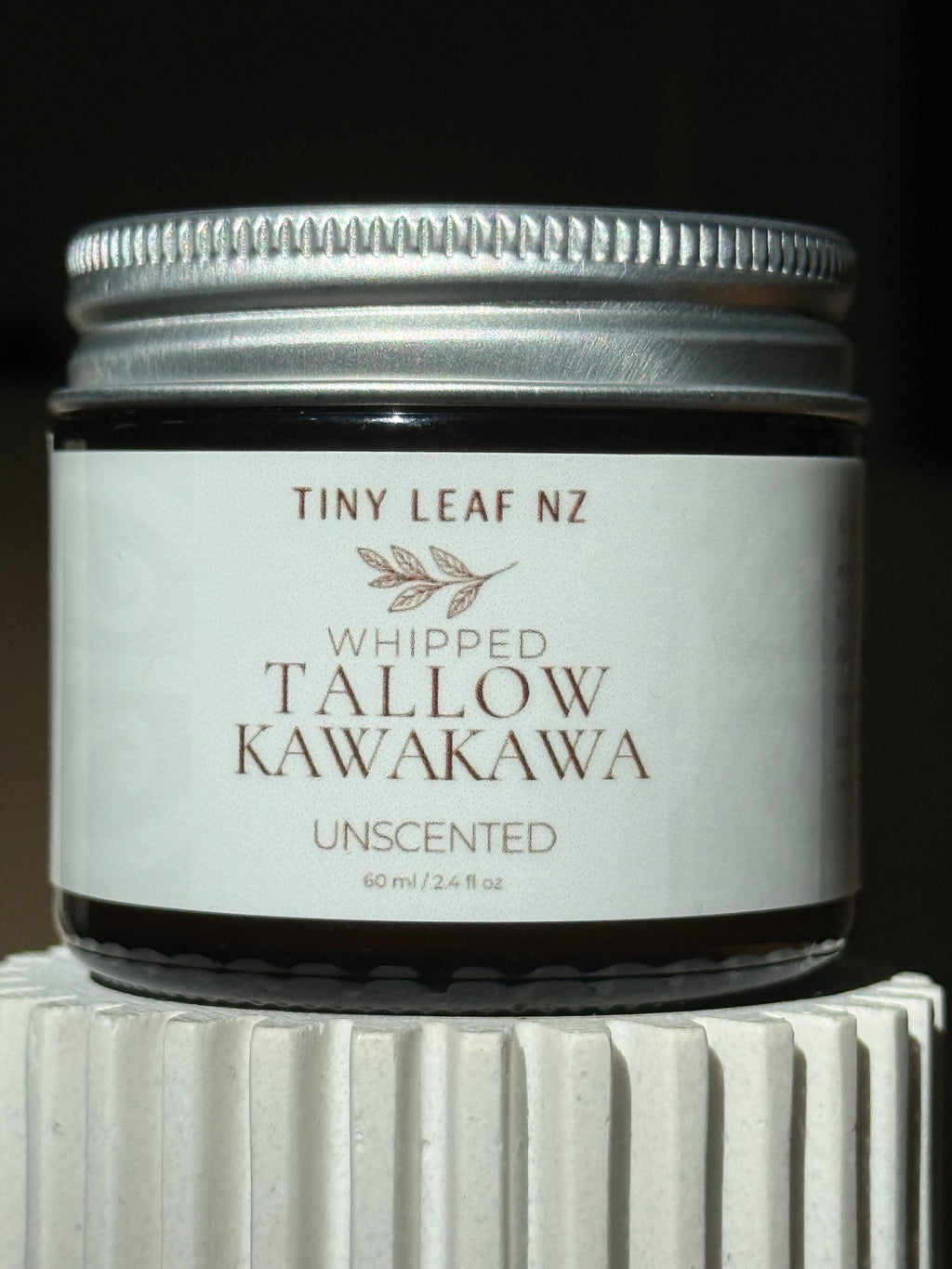 A small glass jar candle with a silver metal lid and a white label reading 'TINY LEAF NZ WHIPPED TALLOW KAWAKAWA UNSCENTED' on a dark background, sitting on a white ribbed pedestal.