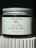 A small glass jar candle with a silver metal lid and a white label reading 'TINY LEAF NZ WHIPPED TALLOW KAWAKAWA UNSCENTED' on a dark background, sitting on a white ribbed pedestal.