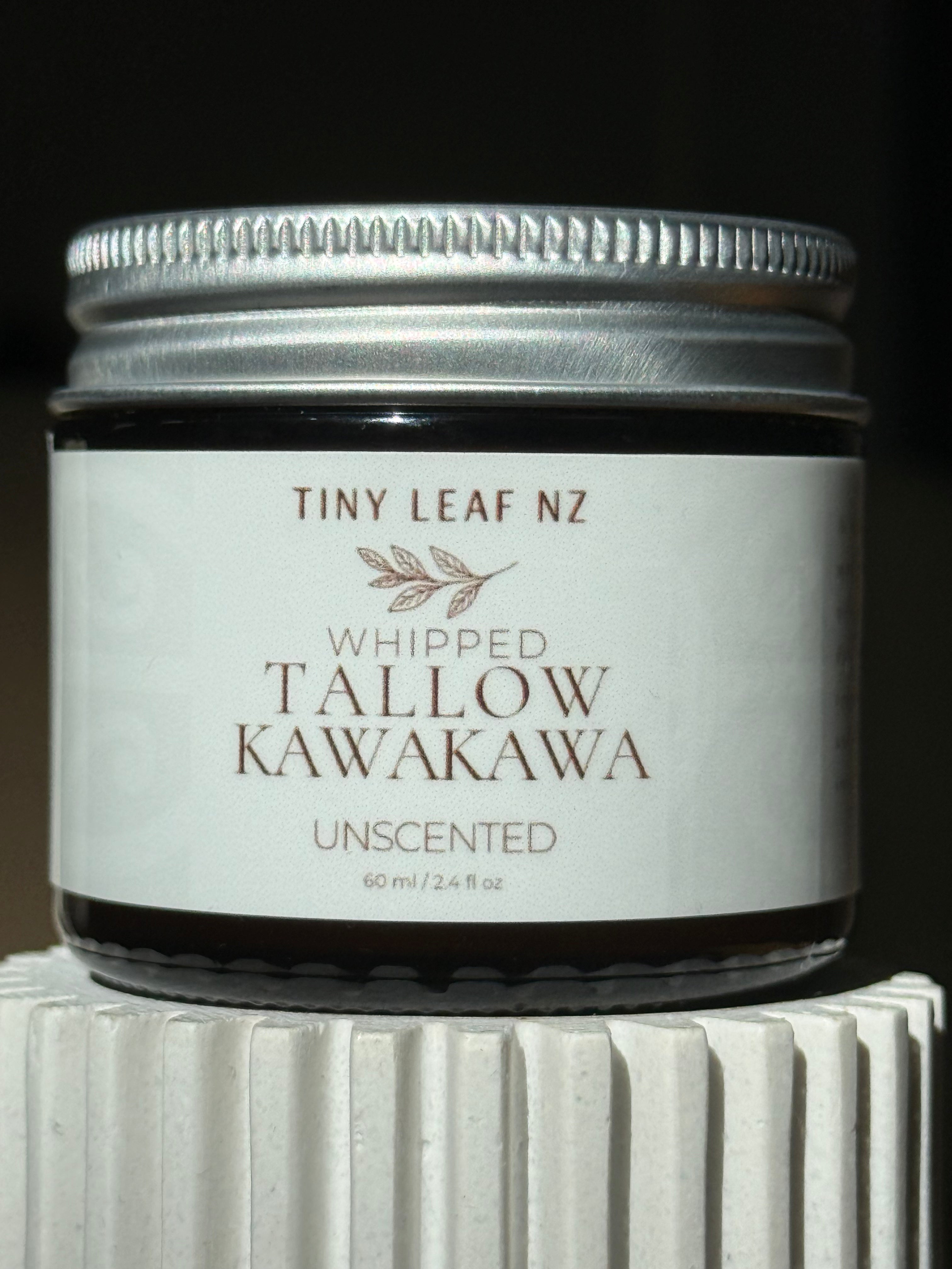 A small glass jar candle with a silver metal lid and a white label reading 'TINY LEAF NZ WHIPPED TALLOW KAWAKAWA UNSCENTED' on a dark background, sitting on a white ribbed pedestal.