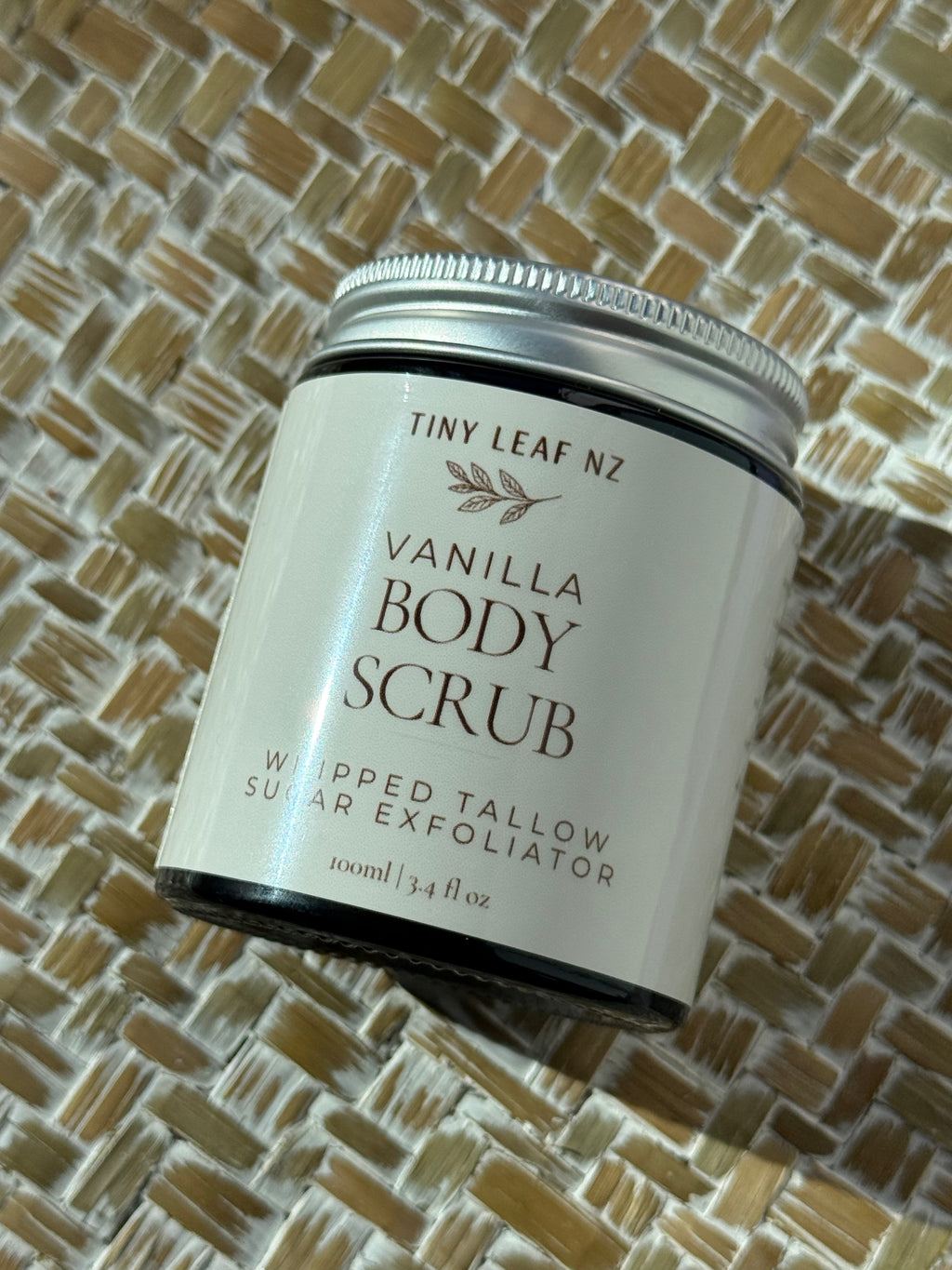 A jar of Tiny Leaf Nz Vanilla Body Scrub, featuring a white label and silver metal lid, resting on a woven straw mat background.