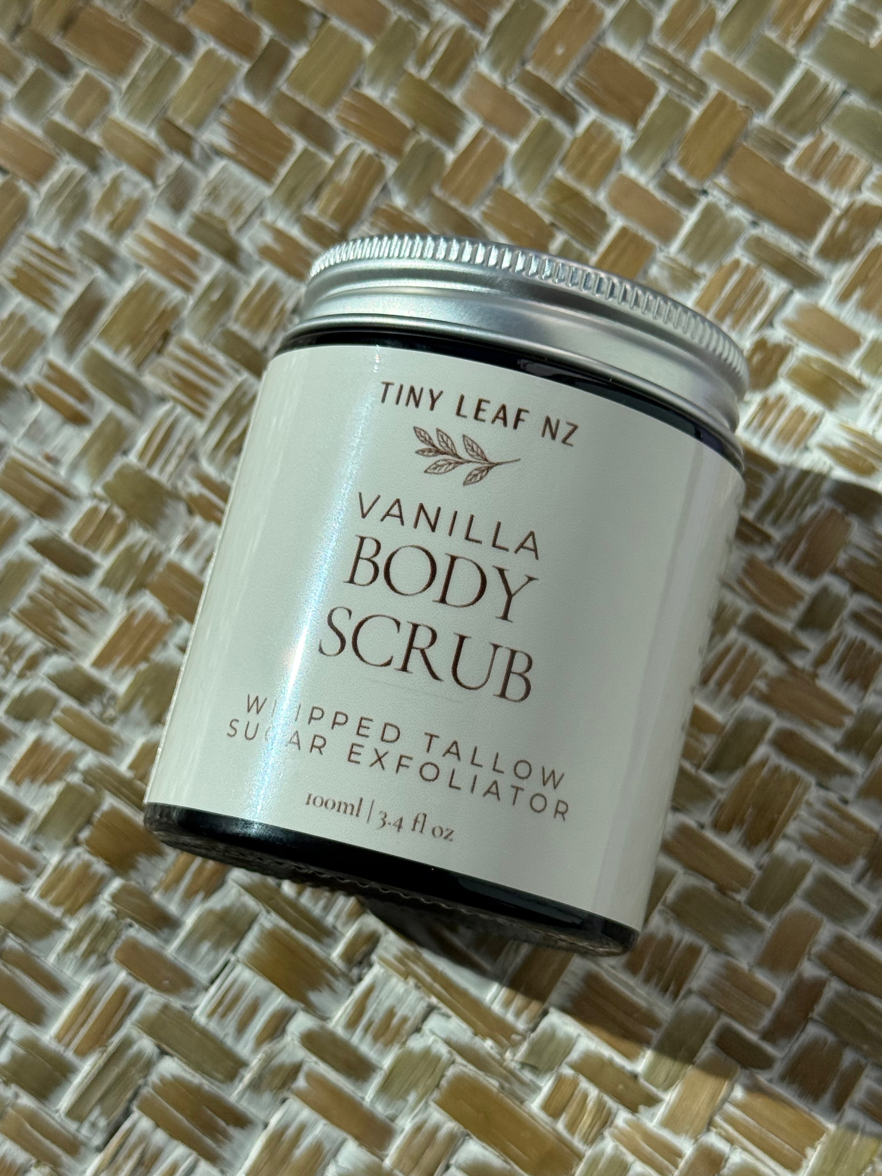 A jar of Tiny Leaf Nz Vanilla Body Scrub, featuring a white label and silver metal lid, resting on a woven straw mat background.