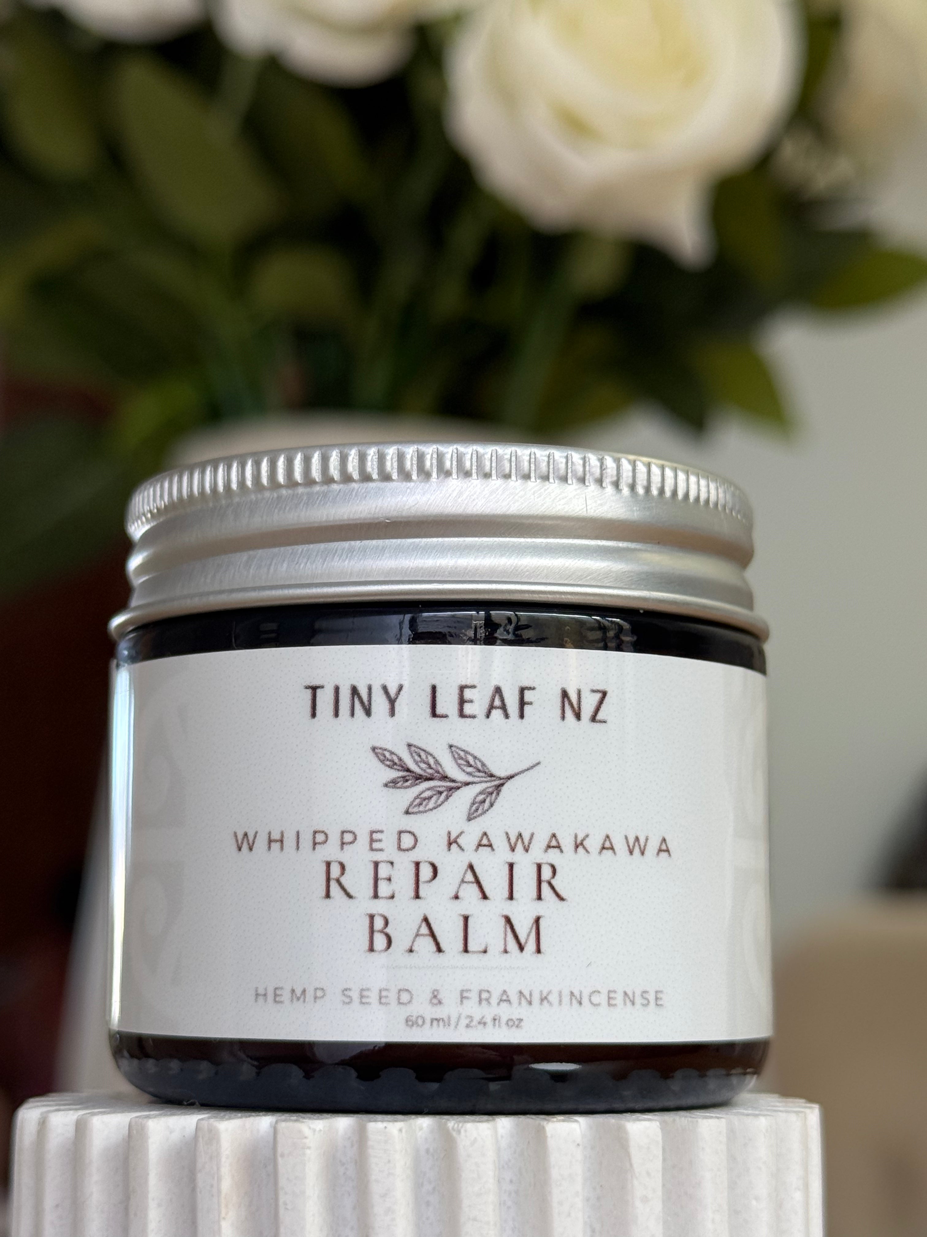 A jar of Tiny Leaf NZ Whipped Kawakawa Repair Balm sits on a white pedestal with a blurred background of white roses and green leaves.