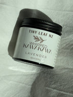 A jar of Tiny Leaf Nz Whipped Kawakawa Lavender, 60ml, with a black lid and white label, resting on a white textured fabric background.