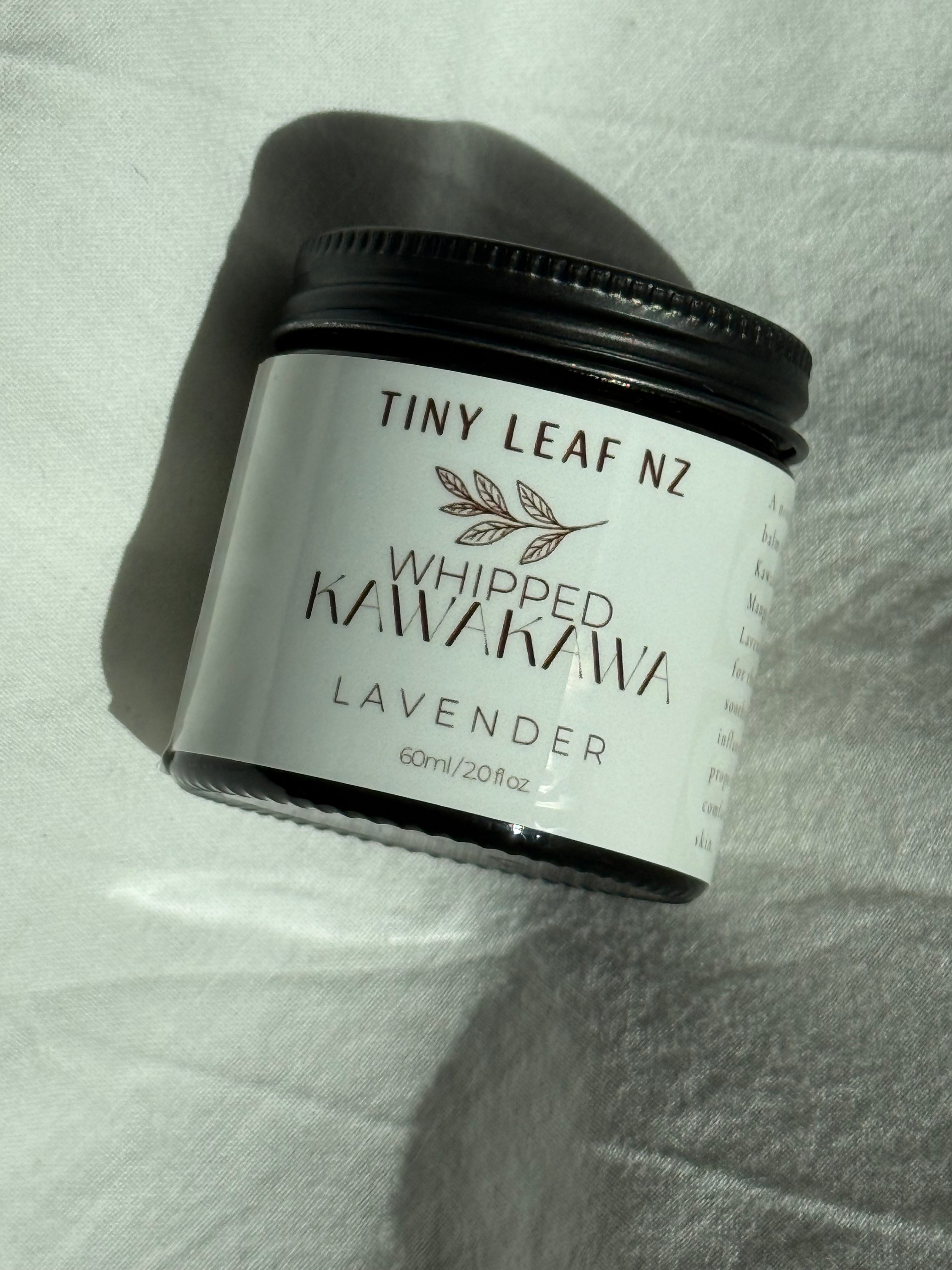 A jar of Tiny Leaf Nz Whipped Kawakawa Lavender, 60ml, with a black lid and white label, resting on a white textured fabric background.