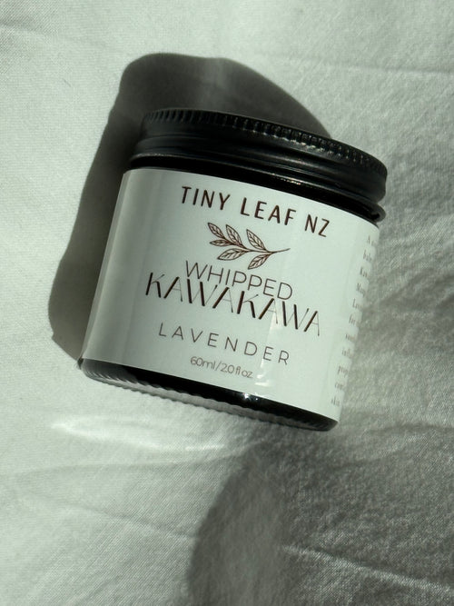 A jar of Tiny Leaf Nz Whipped Kawakawa Lavender, 60ml, with a black lid and white label, resting on a white textured fabric background.