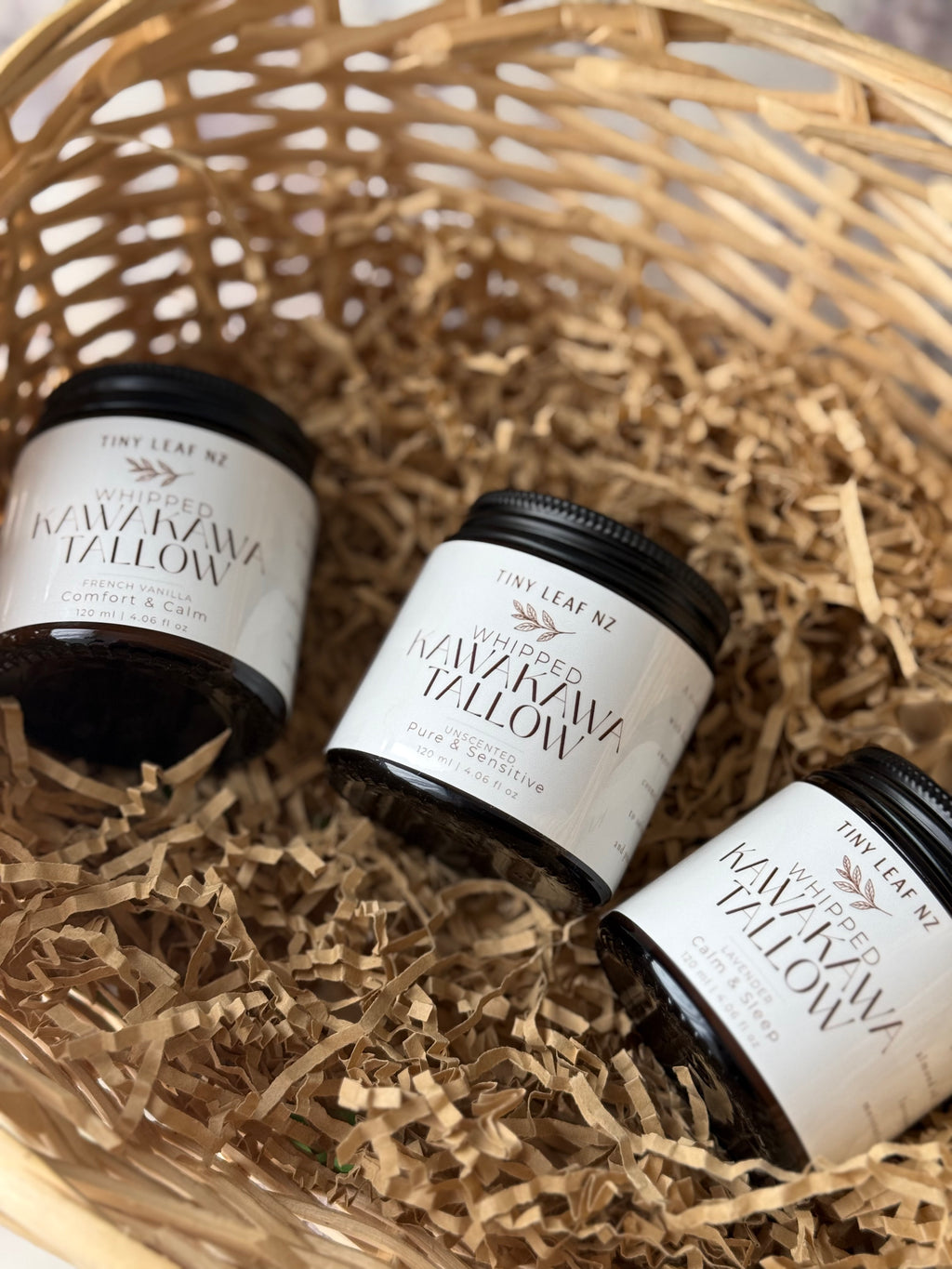 Three small jars of Tiny Leaf NZ Whipped Kawakawa tallow balm arranged in a woven basket with shredded paper filler.