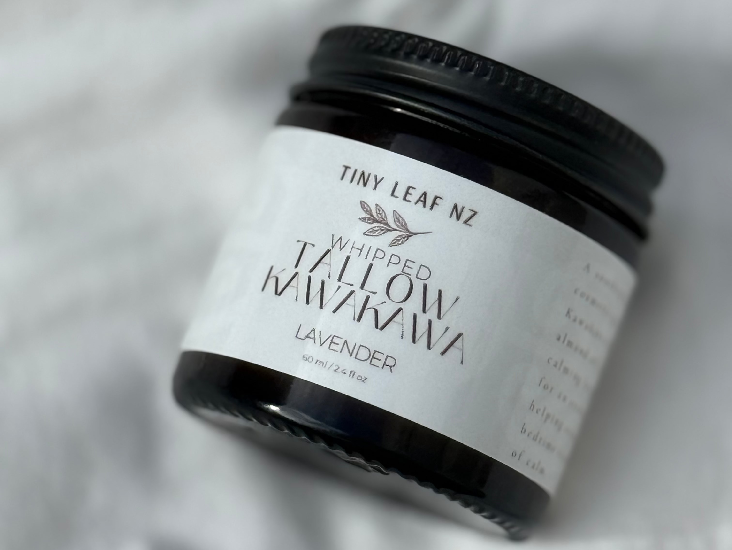 A small black jar of Tiny Leaf NZ 'Whipped Clay Mask' with a white label, resting on a soft, draped white fabric background.