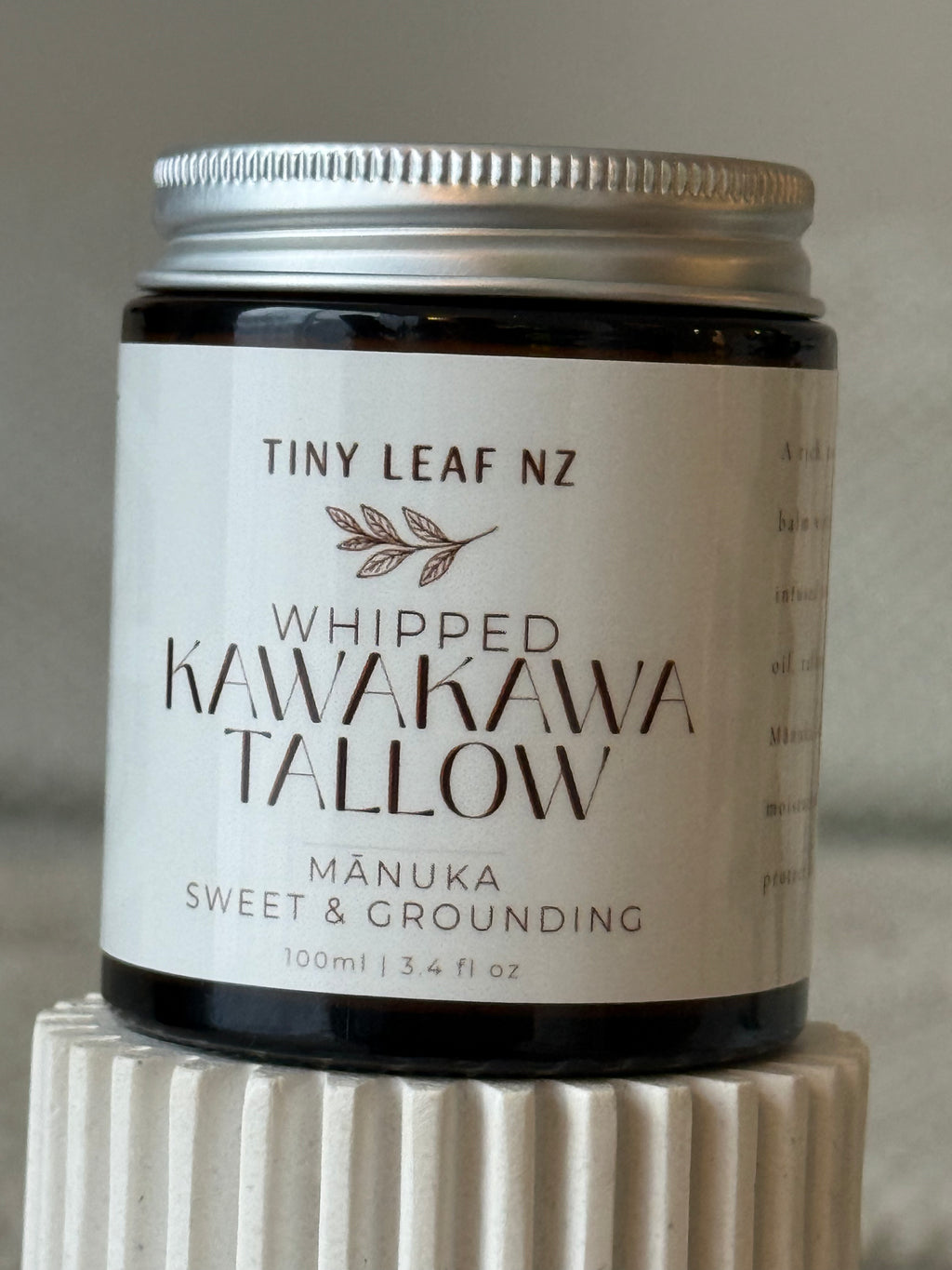 A jar of Tiny Leaf NZ Whipped Kawakawa Tallow (Mâuka Sweet & Grounding) with a white label and silver lid, displayed on a textured white pedestal.