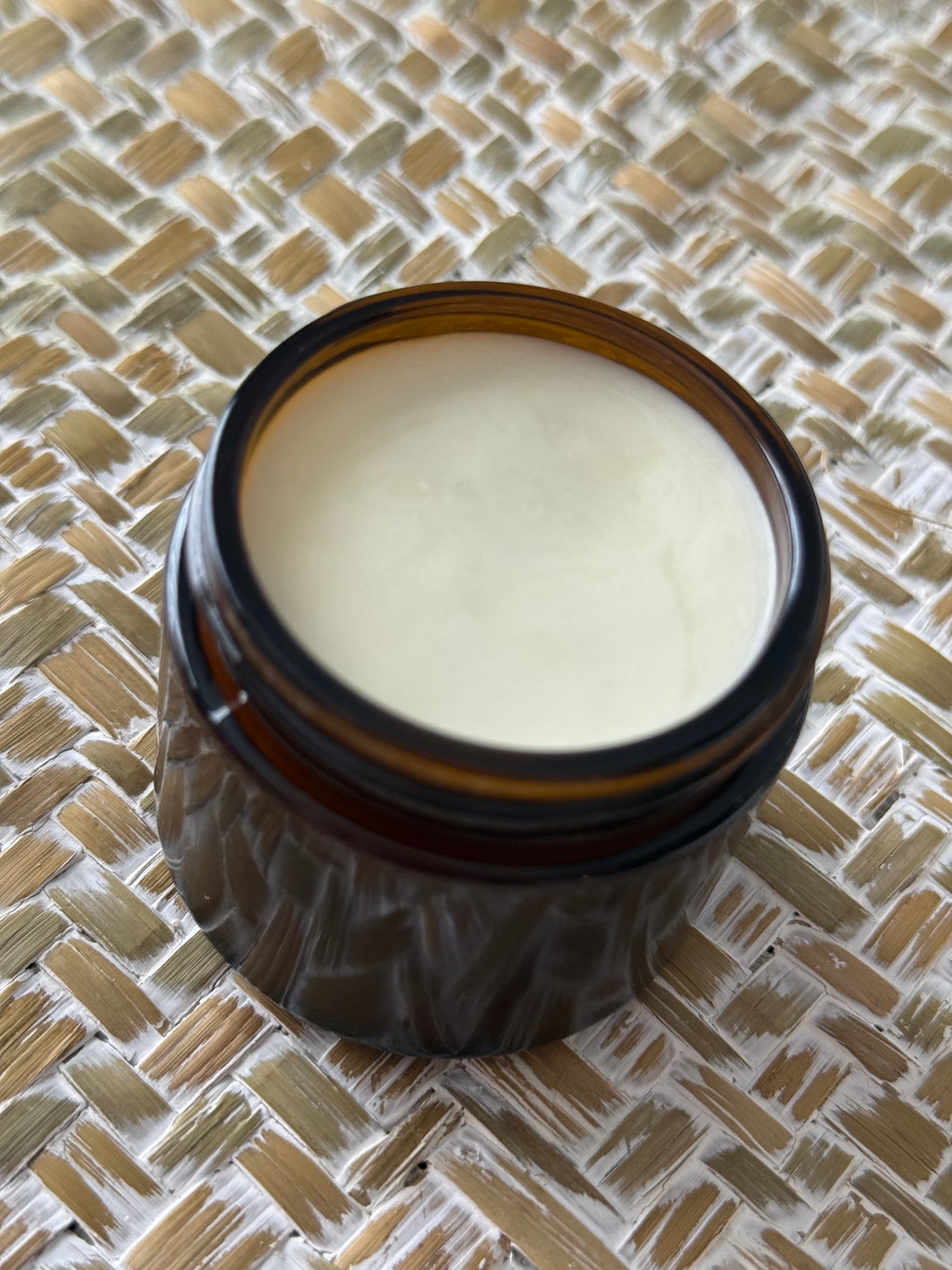 An open amber glass jar filled with a pale cream-colored balm or cream, placed on a woven straw mat background.