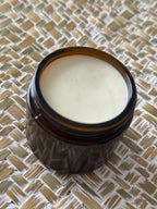 An open amber glass jar filled with a pale cream-colored balm or cream, placed on a woven straw mat background.
