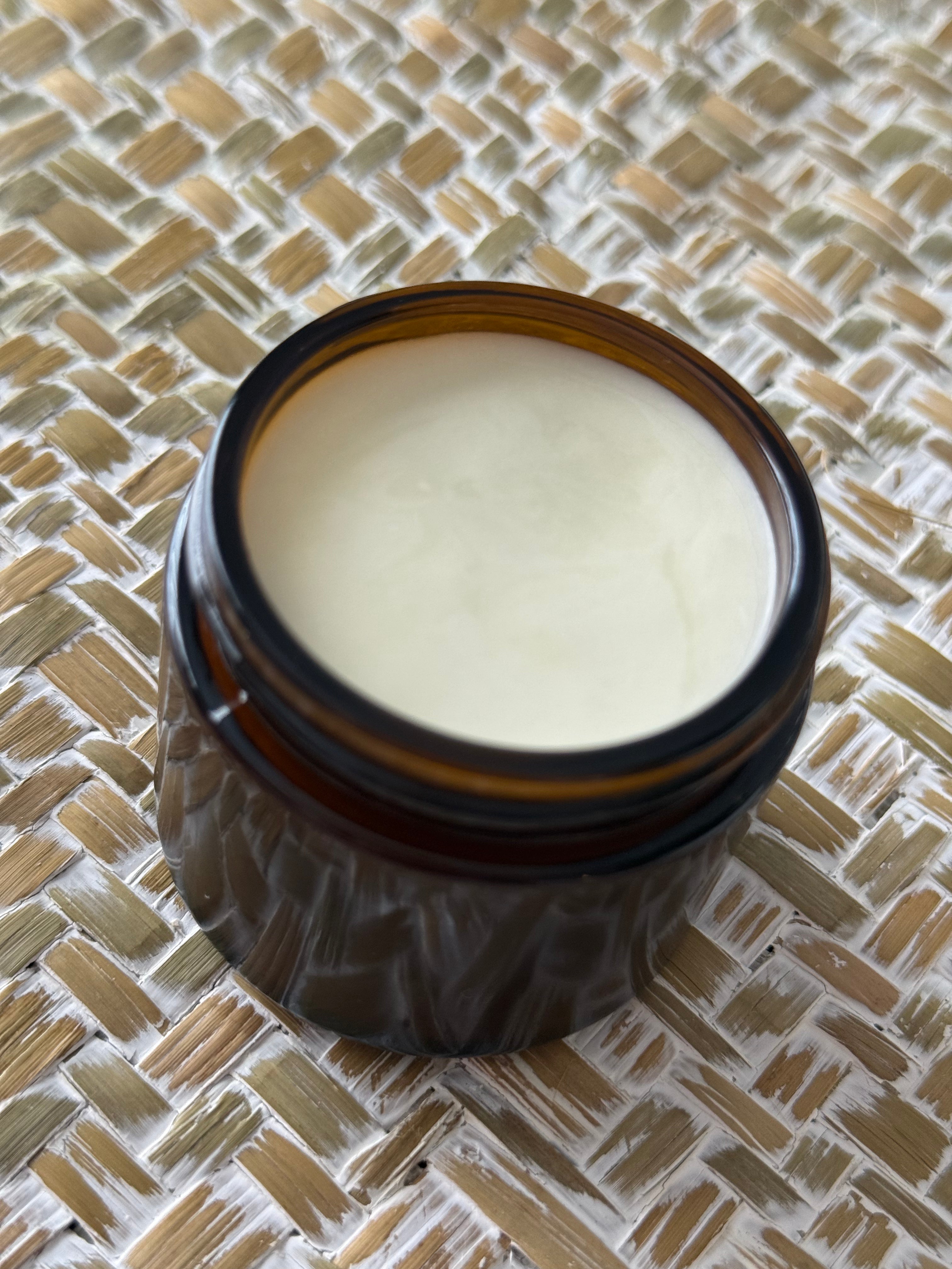 An open amber glass jar filled with a pale cream-colored balm or cream, placed on a woven straw mat background.