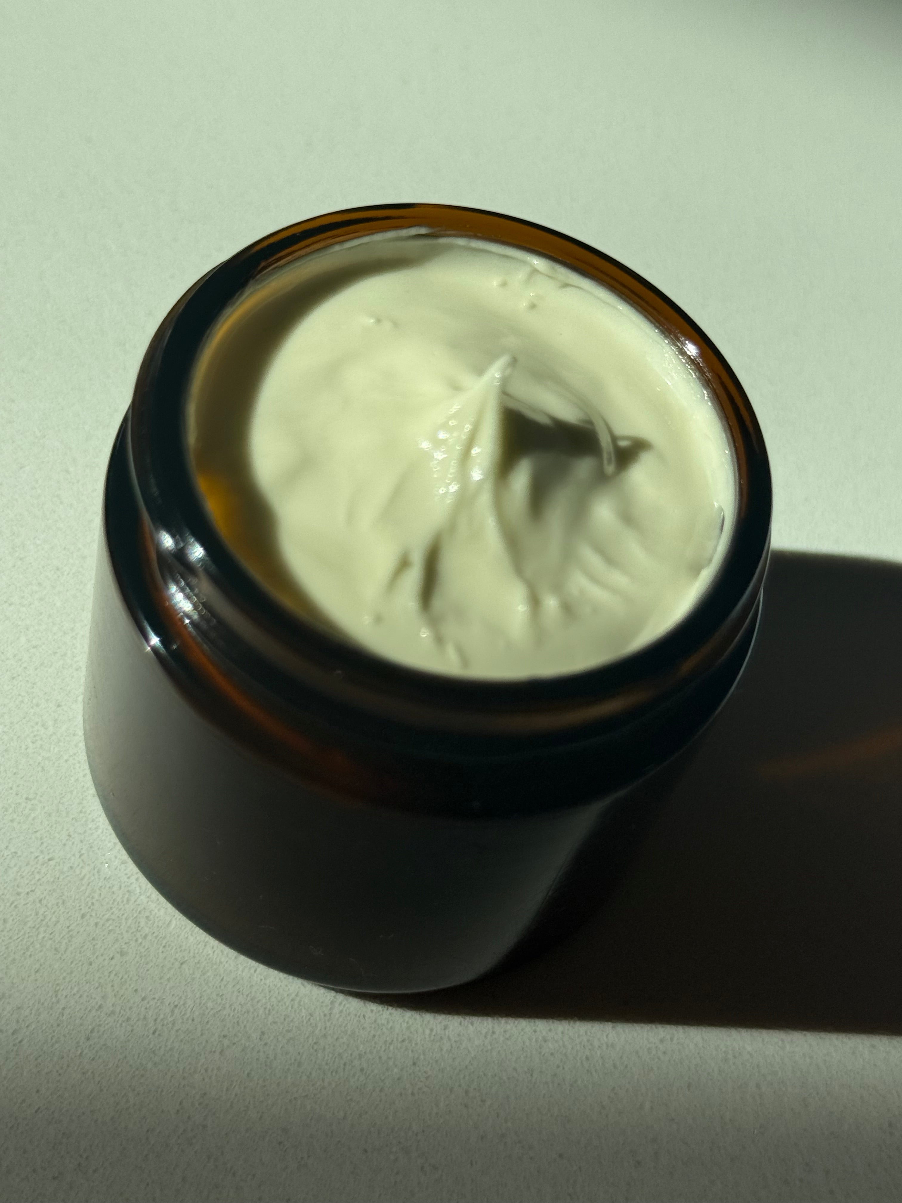 An open amber glass jar filled with thick off-white cream or balm, photographed on a light neutral surface with strong directional sunlight and a hard shadow.