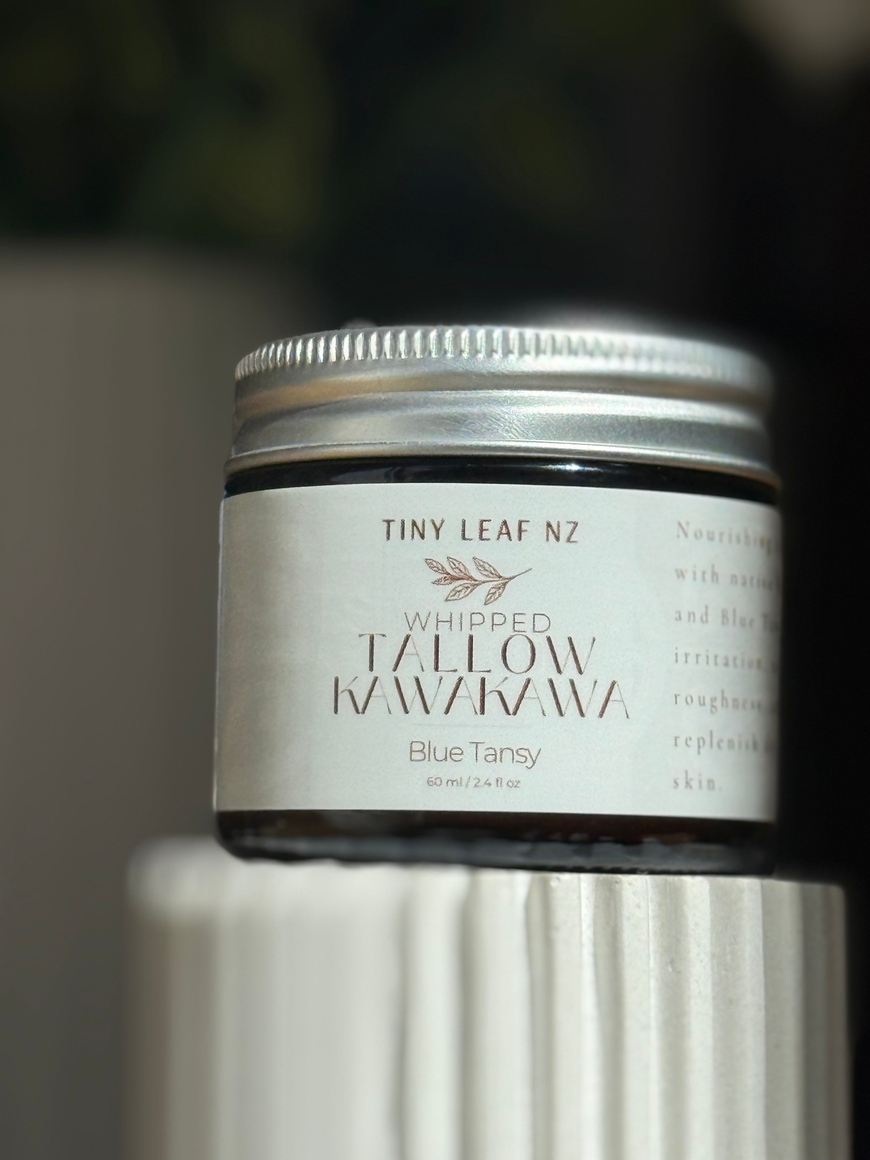 A small cosmetic jar labeled 'TINY LEAF NZ Whipped Tallow Kawakawa Blue Tansy' with a silver lid and white label, photographed close up against a softly blurred dark background.