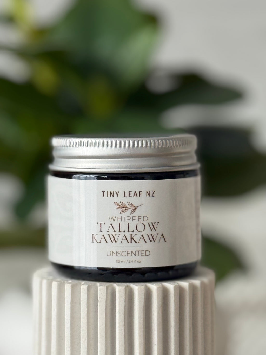 A small tin of Tiny Leaf NZ Whipped Tallow Kawakawa Unscented wax sits on a ribbed white ceramic pedestal with a soft-focus background of green leaves.