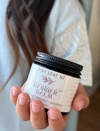 A person holding a small jar of Tiny Leaf NZ Cava Liana + Zinc Barrier Balm. The product is shown in a close-up shot with the person's hand and hair visible in a softly blurred indoor setting.