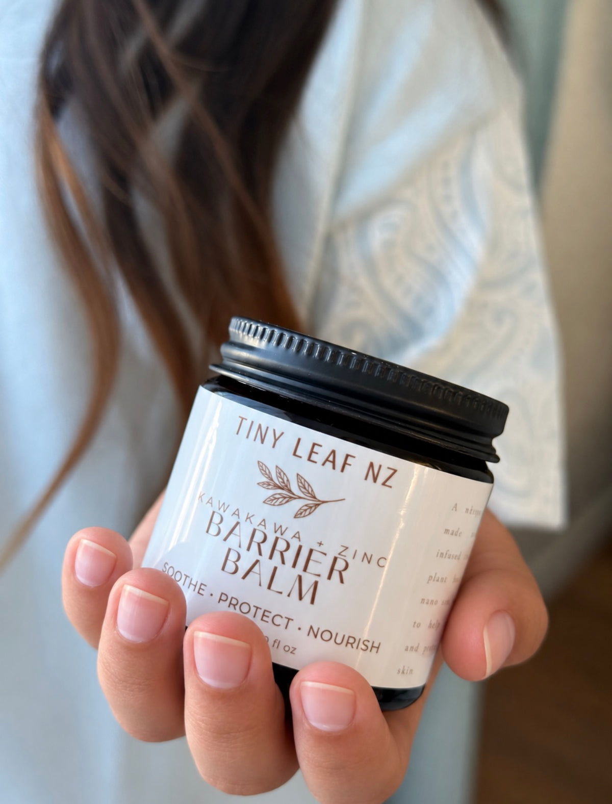 A person holding a small jar of Tiny Leaf NZ Cava Liana + Zinc Barrier Balm. The product is shown in a close-up shot with the person's hand and hair visible in a softly blurred indoor setting.