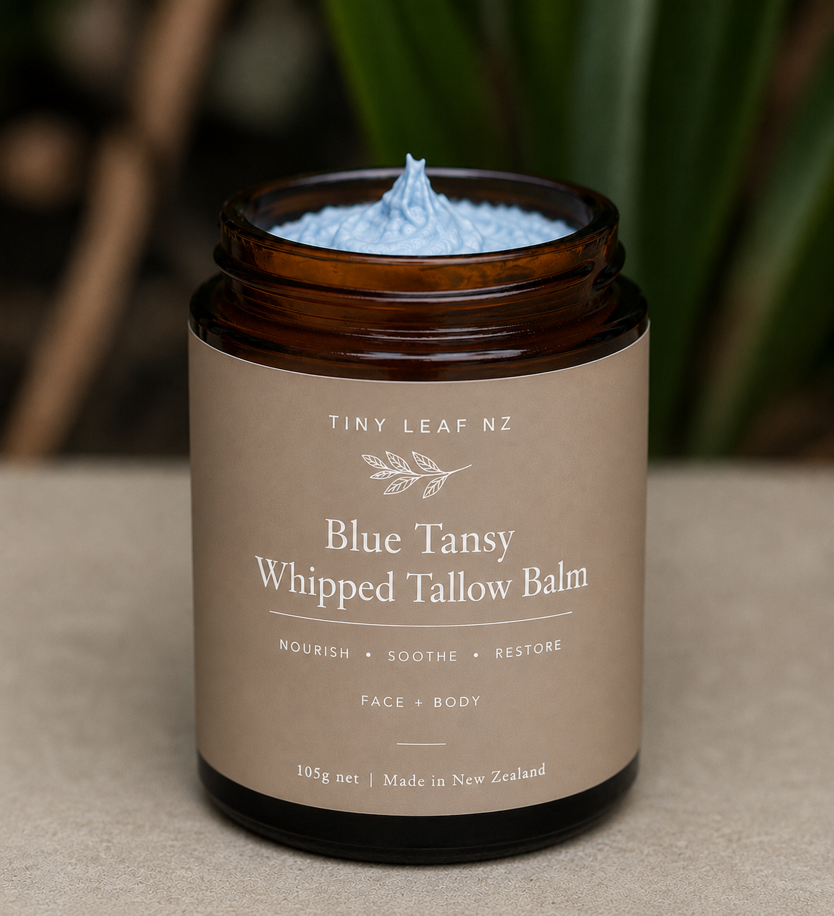 A jar of Tiny Leaf Nz Blue Tansy Whipped Tallow Balm, showing the light blue whipped balm inside an amber glass jar with a beige label, set against a blurred natural background.