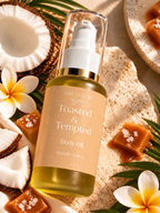 A bottle of Tiny Leaf N2 'Toasted & Tempted' body oil, filled with golden-yellow oil and featuring a beige label with gold accents, styled with tropical props including a halved coconut, white plumeria flowers, caramel candies, and green leaves on a sandy background.