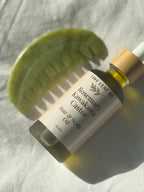 A bottle of Tiny Leaf NZ Rosemary Kawakawa Castor Hair & Scalp Oil with a bamboo dropper cap, styled diagonally next to a green silicone hair comb on a textured white fabric background.