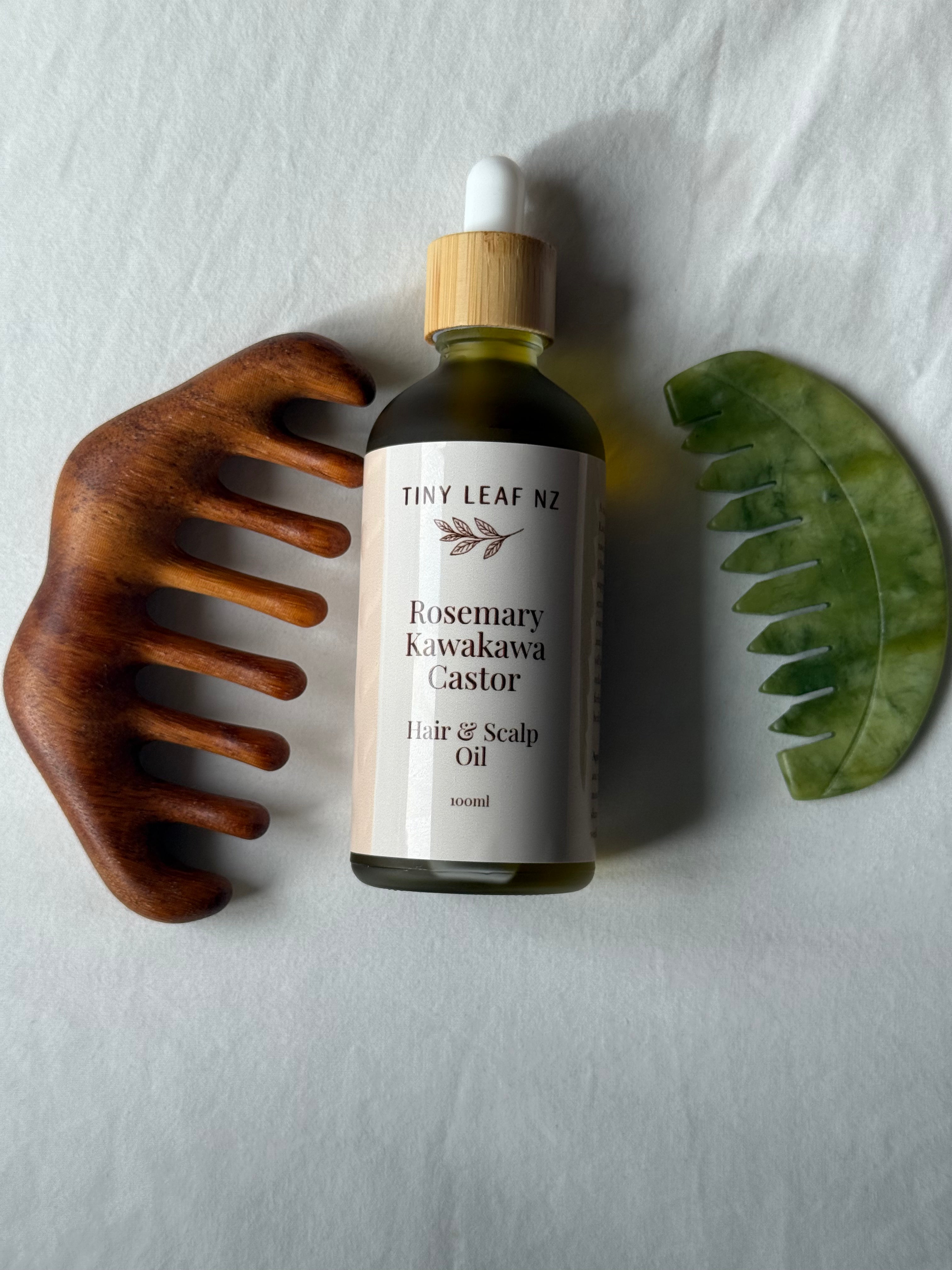 A bottle of Tiny Leaf NZ Rosemary Kawakawa Hair & Scalp Oil is displayed on a textured white background, flanked by a wooden hair comb on the left and a green scalp comb on the right.