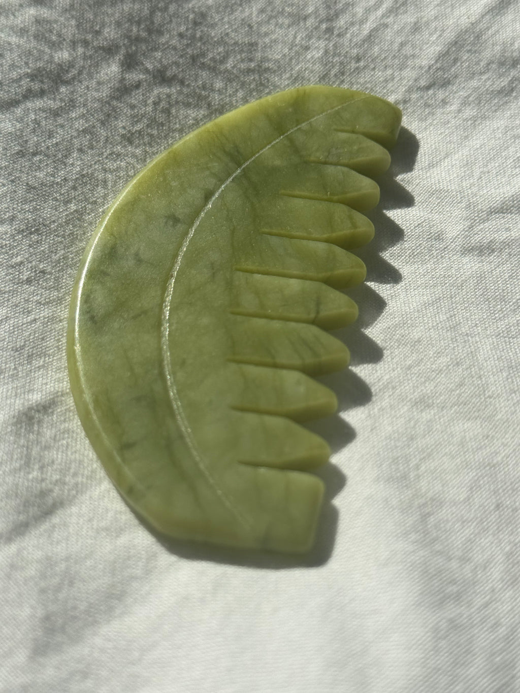 A jade hair comb made of polished green jade, resting on a textured white fabric under direct sunlight.