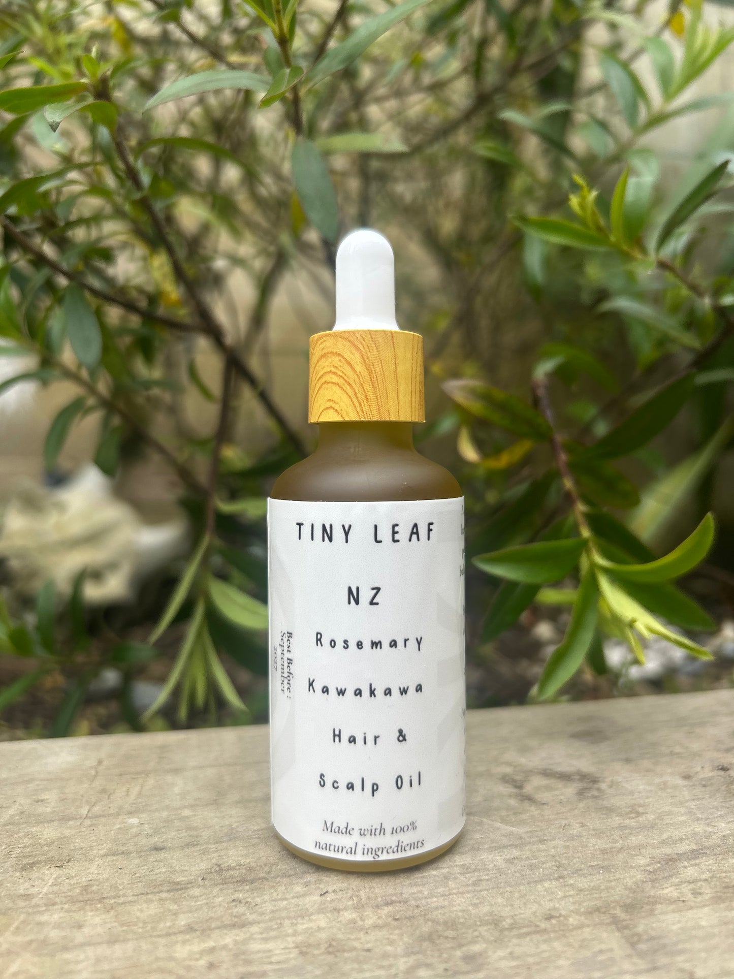 Bottle of Tiny Leaf NZ hair and scalp oil with a dropper on a wooden surface with green foliage in the background.