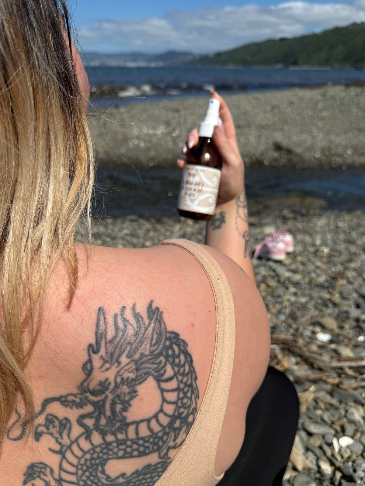 A woman on a rocky beach holds a small amber spray bottle near her shoulder, which features a large black dragon tattoo. The background shows water, distant hills, and sky.