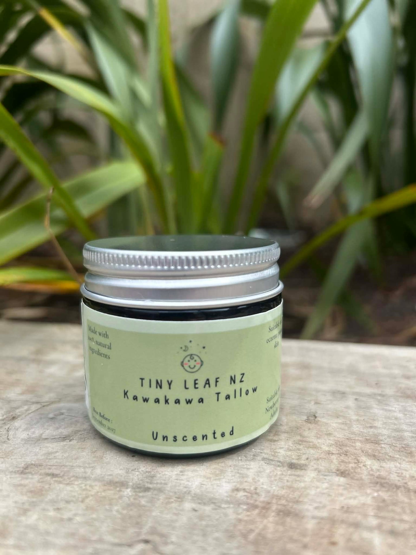 Small green jar with a silver lid on a wooden surface with plants in the background
