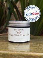 A jar of Tiny Leaf NZ Whipped Kawakawa Tallow (Peach Vanilla) sits on a wooden surface with a green leafy background. An official KidsCan Fundraiser badge is overlaid in the top right corner.