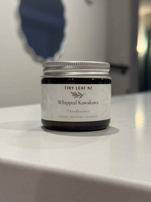 A small glass jar candle labeled 'TINY LEAF NZ Whipped Kawakawa Frankincense' sits on a white countertop with a blurred indoor background.