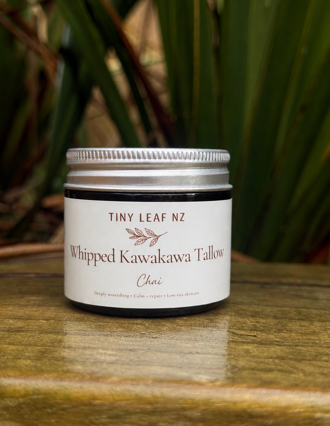 A small jar of Tiny Leaf NZ Whipped Kawakawa Tallow Chai on a wooden surface with a blurred green plant background.